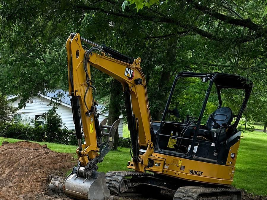 A CAT mini excavator digging into red clay beside a residential lawn.